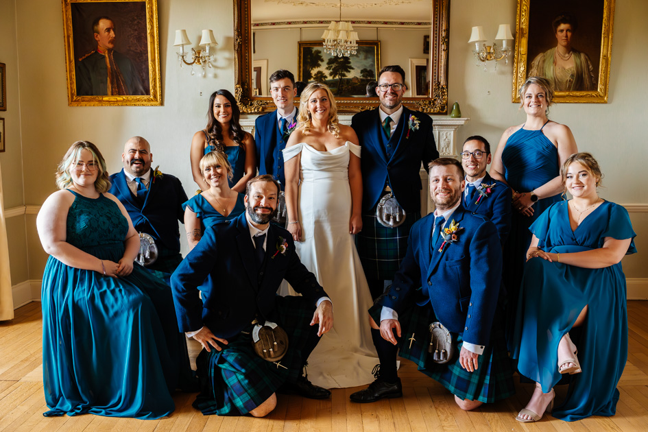 Bridal Party at Colstoun House in Scotland. Bridesmaids wear blue dresses and groomsmen wear blue and green kilts. Bride and groom in the middle