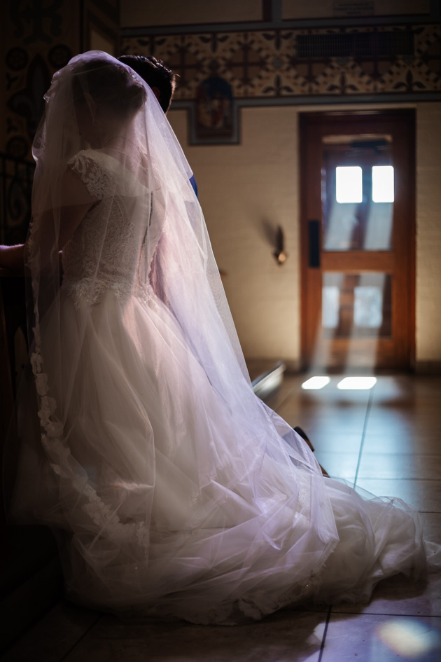 Bride kneels at alter with light from a door in the background illuminating her veil in a church in Arizona